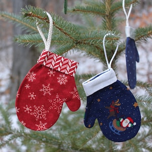 May include: Two Christmas ornaments shaped like mittens. One is red with white snowflakes and a red and white striped cuff. The other is blue with white stars and a white cuff. A reindeer and Santa Claus are on the blue mitten.