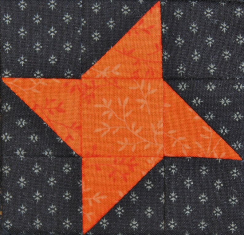 May include: A quilt block with a five-pointed star in orange with a white floral pattern on a black background with a white floral pattern.
