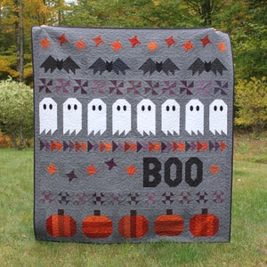 Könnte beinhalten: Eine graue Halloween-Quiltdecke mit orangefarbenen, lila und schwarzen Akzenten. Die Quiltdecke zeigt Fledermäuse, Geister, Sterne, Kürbisse und das Wort "BOO". Die Quiltdecke wird im Freien auf einer Wiese gezeigt.