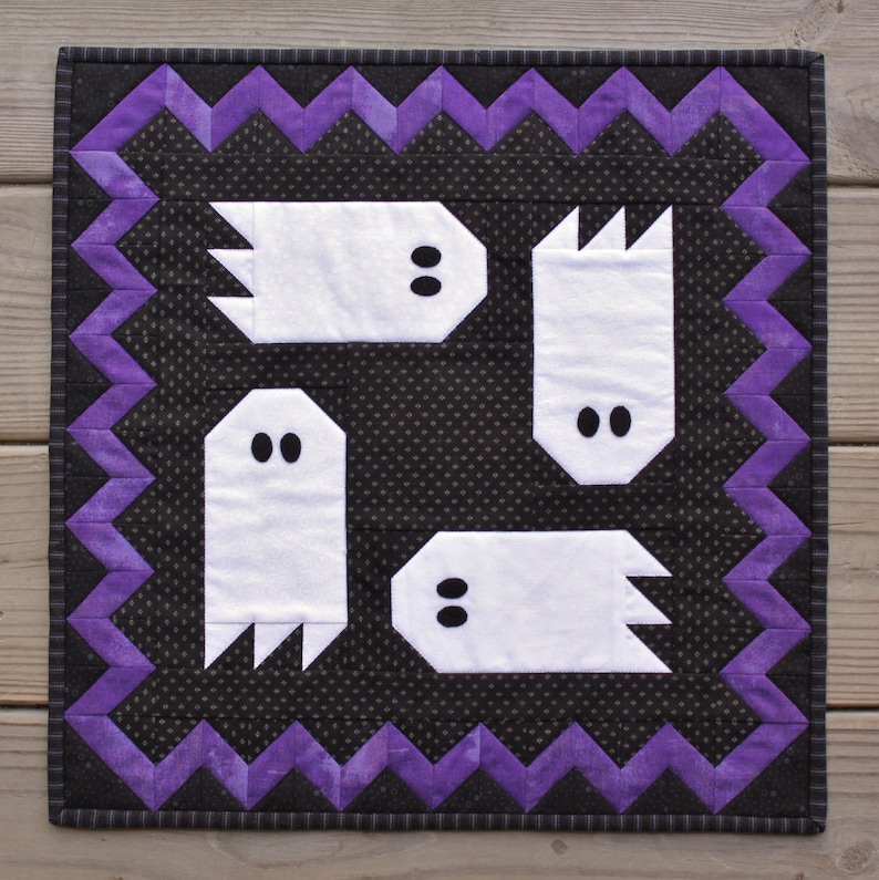 May include: A black and white quilt with a purple zig-zag border. The quilt features four white ghost shapes with black eyes.