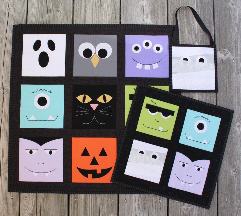 May include: A black Halloween-themed quilt and matching tote bag featuring various monster and character faces. The quilt has a grid pattern with squares of white, gray, purple, and orange. The tote bag has a black handle.
