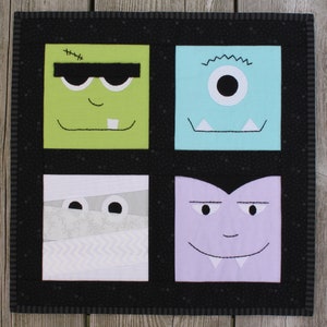May include: A square Halloween-themed quilt featuring four colorful monster faces. Each face is a different color: green, blue, white, and purple. The quilt has a black border with a dotted pattern and a striped edge.