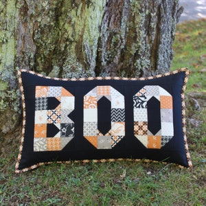 May include: A black pillow with a white and orange patchwork design spelling out "BOO". The pillow is sitting on a bed of green grass.