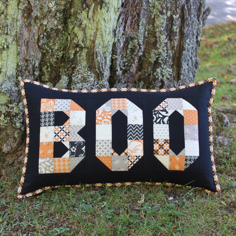 May include: A black pillow with a white and orange patchwork design spelling out "BOO". The pillow is sitting on a bed of green grass.