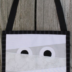 May include: A handmade Halloween-themed tote bag featuring a mummy design. The bag has a black strap and a black and white plaid border. The mummy's face is created with white fabric strips and large black eye cutouts.