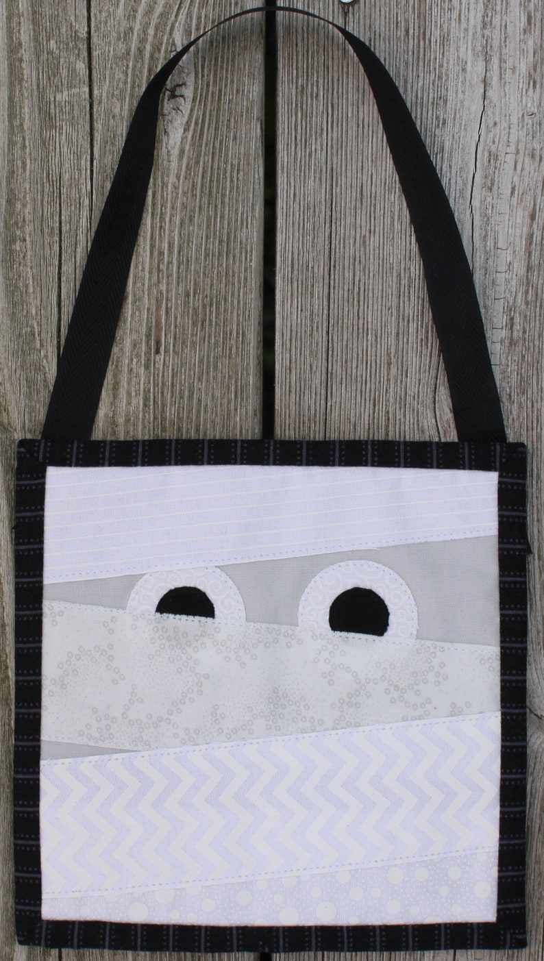 May include: A handmade Halloween-themed tote bag featuring a mummy design. The bag has a black strap and a black and white plaid border. The mummy's face is created with white fabric strips and large black eye cutouts.