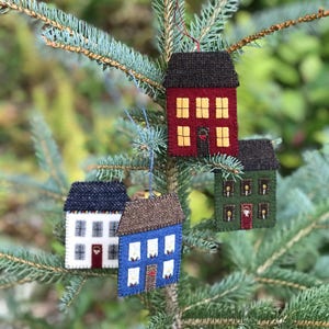 May include: Four felt house ornaments hanging on a pine tree branch. The houses are red, green, blue, and white with brown roofs. Each house has windows and a door.