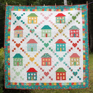 May include: A white quilt with a patchwork design featuring colorful houses and hearts. The quilt is bordered with a teal and red patterned fabric.