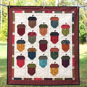 Puede incluir: Un edredón hecho a mano con un patrón repetido de formas de bellotas coloridas. El edredón tiene un fondo blanco con un borde rojo y está colgado al aire libre en un tendedero. Las bellotas son de varios colores, incluyendo marrón, rojo y verde.