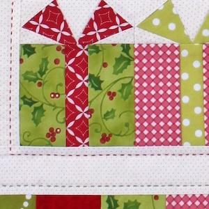 May include: Close-up of a Christmas-themed quilt block featuring gift box designs. The boxes are made of green fabric with holly patterns and red fabric with white polka dots and diamond patterns. White bows and ribbons complete the festive look.
