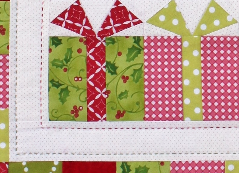 May include: Close-up of a Christmas-themed quilt block featuring gift box designs. The boxes are made of green fabric with holly patterns and red fabric with white polka dots and diamond patterns. White bows and ribbons complete the festive look.