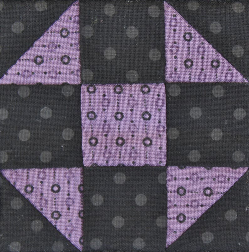 May include: A quilt block with a purple and black geometric pattern. The block is made up of four squares, each with a different pattern. The squares are arranged in a checkerboard pattern.
