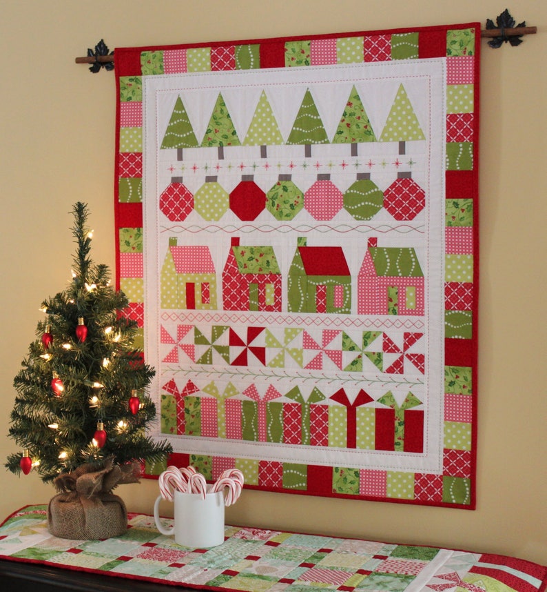 May include: A Christmas-themed wall hanging with a red and green patchwork border. The central design features appliqued Christmas trees, ornaments, houses, pinwheels, and wrapped gifts. A small Christmas tree with lights and a mug of candy canes are in the foreground.