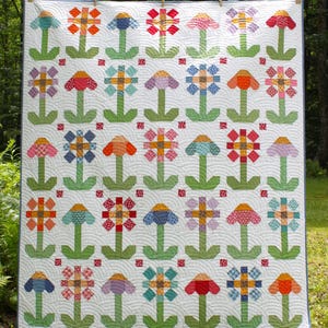 May include: A colourful handmade quilt with a repeating floral pattern. The flowers are in shades of red, orange, blue, and purple, with green stems and leaves. The quilt is displayed outdoors, highlighting the detailed patchwork design.