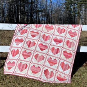 May include: A pink and white quilt with a pattern of hearts. The quilt is made of fabric scraps and is quilted with a white thread. The quilt is hanging on a white fence.