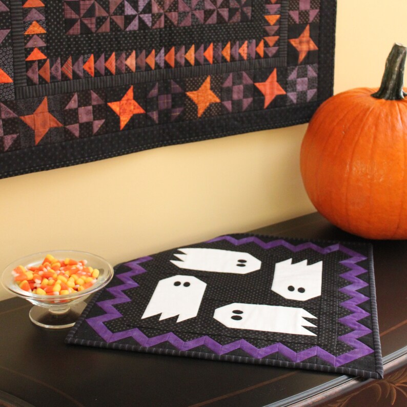 May include: A black and purple quilted table runner with a white ghost design. The runner is on a dark wood table with a bowl of candy corn and a large orange pumpkin.