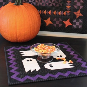 May include: A black and purple quilted table runner with a zigzag border features white ghost appliques. A bowl of candy corn sits on the runner, and a large orange pumpkin is in the background.