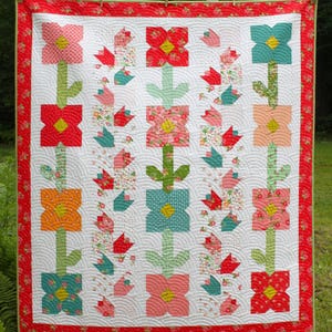 May include: A handmade quilt with a floral pattern. The quilt displays large, stylised flowers in pink, red, orange, and teal, set against a white background. The quilt is bordered with a red floral pattern and measures approximately 152 cm by 178 cm.