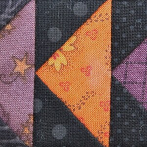 May include: A patchwork quilt block with a variety of fabrics in orange, black, and purple. The block is made up of triangles and squares, and the fabrics have different patterns, including polka dots, floral designs, and geometric shapes.