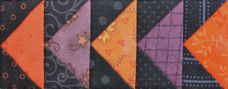 May include: A patchwork quilt block with a variety of fabrics in orange, black, and purple. The block is made up of triangles and squares, and the fabrics have different patterns, including polka dots, floral designs, and geometric shapes.