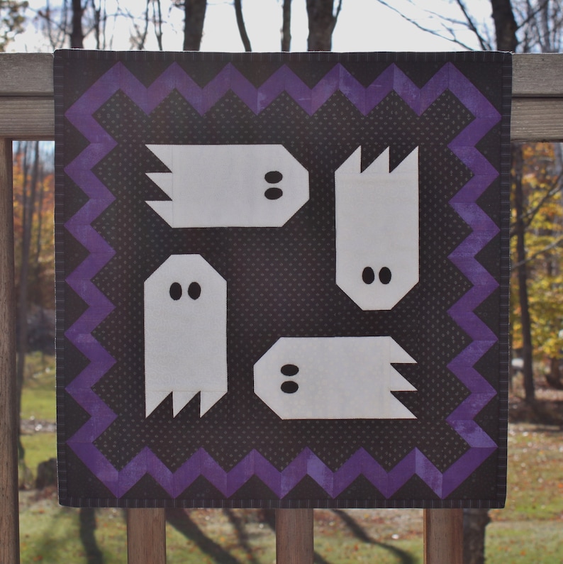 May include: A black and white quilt with a purple border. The quilt features four white ghost shapes with black eyes. The quilt is perfect for Halloween decor.