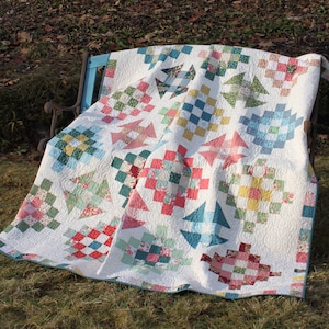 May include: A white quilt with a patchwork design featuring squares, stars, and other geometric shapes in shades of pink, blue, green, and yellow. The quilt is folded and draped over a blue bench.