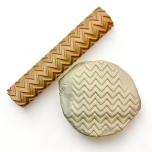 May include: A tan, textured rolling pin with a chevron pattern next to a piece of clay with the same pattern pressed into it. The rolling pin is cylindrical and the clay is a light green color.