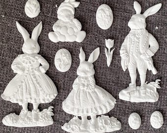 May include: A collection of white Easter-themed decorations, including several bunny figurines in various poses, Easter eggs with floral designs, and a tulip. The figurines are detailed and appear to be made of plaster or a similar material.