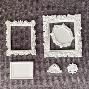 May include: A collection of white ornate picture frames and decorative elements. The frames vary in size and shape, including rectangular and oval designs. The decorative elements include a small fan-shaped piece and a circular piece.