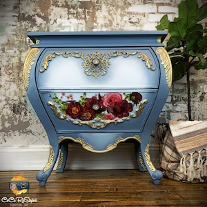 A 2 drawer end table that is blended blue and white. On the bottom drawer, there are various roses. Along the sides are ornate details painted gold. In the bottom left corner, it reads CeCe Restyled furniture art.