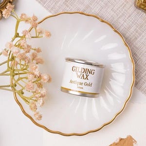 May include: A small can of "Gilding Wax Antique Gold" sits on a white, scalloped-edge plate with a gold rim. The can has a silver lid and a white label with gold lettering. A sprig of pink flowers is to the left of the plate.