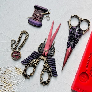 May include: A set of three decorative scissors with ornate designs. The scissors are made of metal and have a purple and gold finish. One pair of scissors has a red and gold finish. The scissors are displayed on a white surface with a red and white compass.