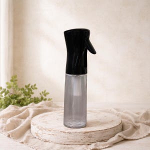 May include: A clear, cylindrical spray bottle with a black top and trigger. The bottle is empty and sits on a round, light-colored wooden stand. The background is a neutral beige with a hint of green foliage.