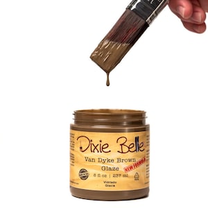 May include: A brown paint glaze in a jar with the label "Dixie Belle Van Dyke Brown Glaze New Formula 8 fl oz | 237 ml Vidriado Glacis". A paintbrush with brown paint is dripping onto the jar.