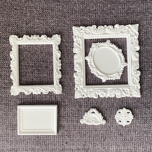 May include: A collection of white ornate picture frames and decorative elements. The frames vary in size and shape, including rectangular and oval designs. The decorative elements include a small fan-shaped piece and a circular piece.
