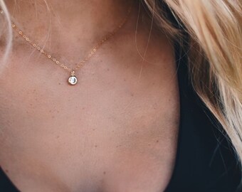 May include: A delicate gold necklace with a small, round, clear gemstone pendant. The necklace is worn against a person's skin, with blonde hair visible in the background. The pendant is the focal point of the image.
