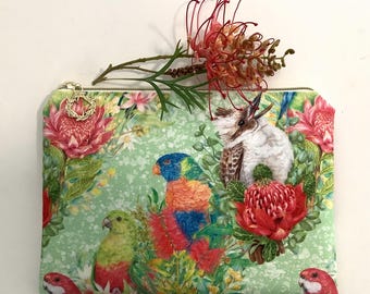 Puede incluir: Un estuche rectangular con cremallera que presenta un fondo verde claro con un estampado de loros coloridos, cucaburras y flores de protea rojas. El estuche tiene un tirador de cremallera de metal dorado con un dije en forma de corona.