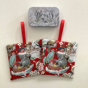 May include: Two red fabric sachets with a whimsical print featuring koalas in Santa hats, sailing in boats, and other characters. A silver tin with lavender sprigs and the word "Lavender" is above the sachets. Red ribbon loops are attached to the sachets.