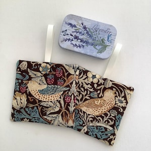 May include: A brown fabric pouch with a floral and bird design. The pouch has two white straps and is closed. A small tin box with a lavender design and the word "Lavender" printed on it is in the background.