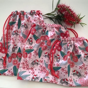 May include: Three drawstring bags with a pink floral and mouse print. The bags have red drawstrings and are decorated with red flowers, green leaves, and grey mice wearing Santa hats. The bags are in different sizes.