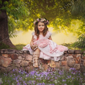 May include: A young girl wearing a pink dress and a flower crown sits on a stone wall. She is surrounded by purple flowers and green foliage.