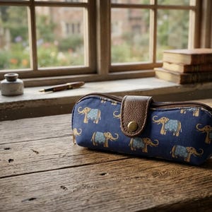 Puede incluir: Un estuche para gafas azul marino con un estampado repetido de elefantes en beige y azul claro. El estuche tiene una solapa de cuero marrón con un cierre de botón dorado. Está sobre una superficie de madera.
