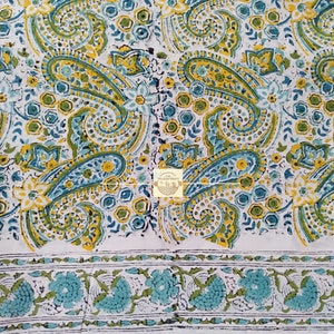 Könnte beinhalten: Weißer Stoff mit einem sich wiederholenden Paisley-Muster in Gelb, Türkis und Grün. Das Design umfasst florale Elemente und einen Rand aus türkisfarbenen Blumen und grünen Blättern. Der Stoff hat einen handgefertigten Look.