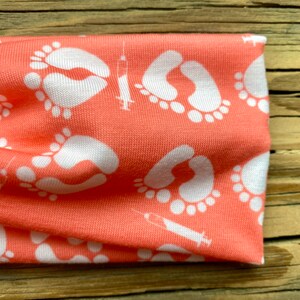 May include: Coral-colored headband featuring a repeating pattern of white baby footprints and syringes. The fabric is soft and appears to be a knit material. The headband is designed for comfort and style.