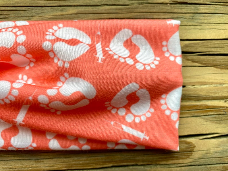 May include: Coral-colored headband featuring a repeating pattern of white baby footprints and syringes. The fabric is soft and appears to be a knit material. The headband is designed for comfort and style.