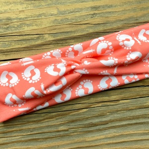 May include: Coral-colored headband with a white pattern of baby footprints and syringes. The twisted fabric headband is designed for comfort and style. This accessory is perfect for those who want to celebrate or support new beginnings.