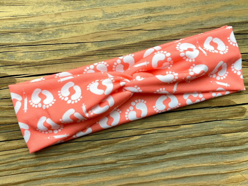 May include: Coral-colored headband with a white pattern of baby footprints and syringes. The twisted fabric headband is designed for comfort and style. This accessory is perfect for those who want to celebrate or support new beginnings.