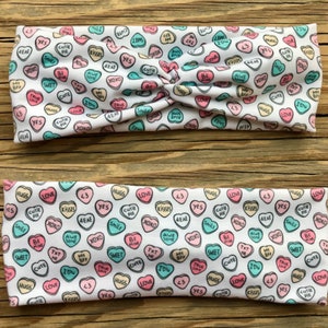 May include: Two white headbands with a repeating pattern of colorful candy hearts. Each heart has a different message, such as "Kisses," "Cute," and "Be Mine." One headband has a twisted knot.