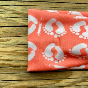 May include: Coral-colored headband featuring a repeating pattern of white baby footprints and syringes. The fabric is soft and stretchy, ideal for comfort. This headband is a stylish accessory for expecting parents or those celebrating new life.