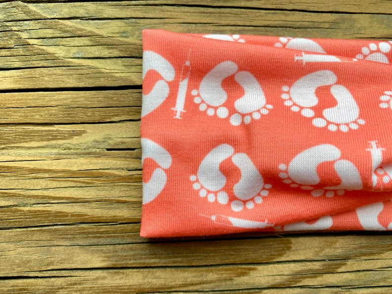May include: Coral-colored headband featuring a repeating pattern of white baby footprints and syringes. The fabric is soft and stretchy, ideal for comfort. This headband is a stylish accessory for expecting parents or those celebrating new life.
